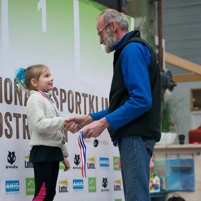 Een oudere man reikt glimlachend een medaille uit aan een jong meisje met een paardenstaart, voor een banner van een sportevenement.